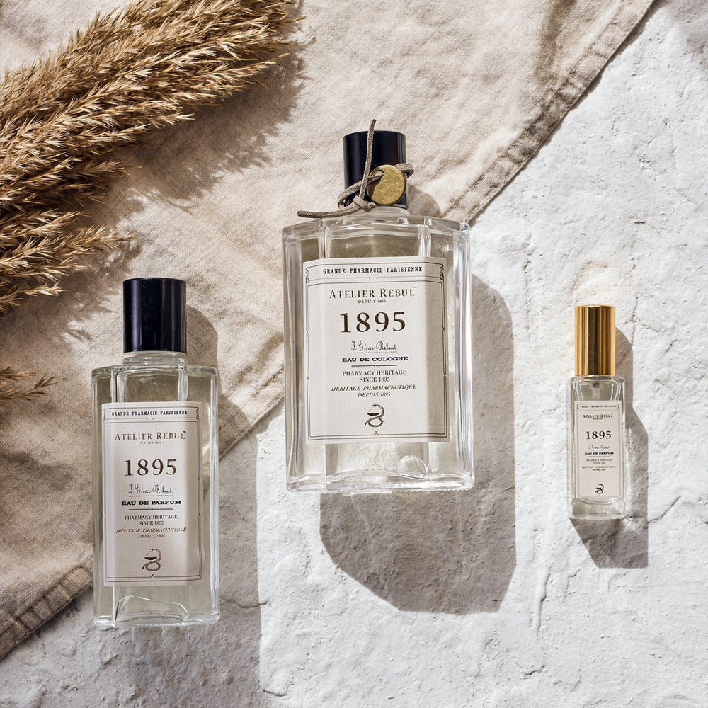 Assortiment van parfums met de 1895 geur van Atelier Rebul. op de foto liggen een kleine parfum, een grote parfum en een eau de cologne. De achtergrond is een beige textiel doek en wit pleisterwerk. De foto werd in bovenaanzicht gemaakt.