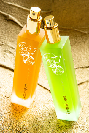 Fixerende sprays van i.am.klean in het oranje en groen. De oranje fixing spray heeft een glanzend effect voor droge huid, de groene een matterend effect voor vette huid. De flessen staan op een achtergrond van zand.