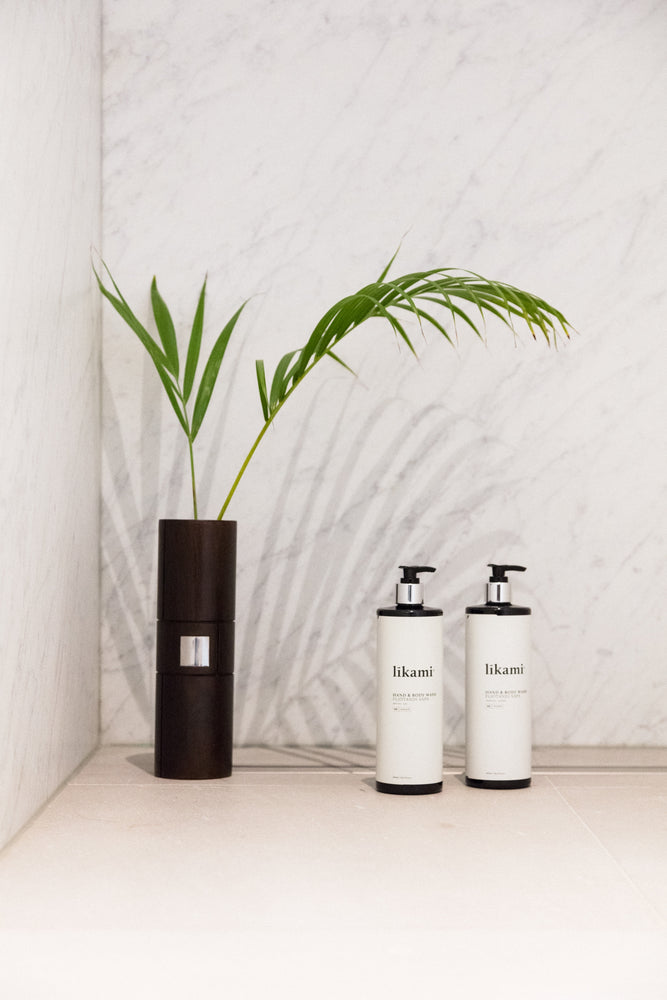 Twee grote douchegels van Likami voor een marmeren wand. De lichaamsproducten staan naast een groen palmblad. De producten zitten in een zwarte fles met pomp en wit etiket.