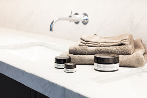 Assortiment aan lichaamsverzorging van Likami. De producten staan naast een marmeren lavabo met bruine handdoeken in een badkamer. Op de foto staan de natuurlijke body crème, lippenbalsem en hydraterende dagcrème.