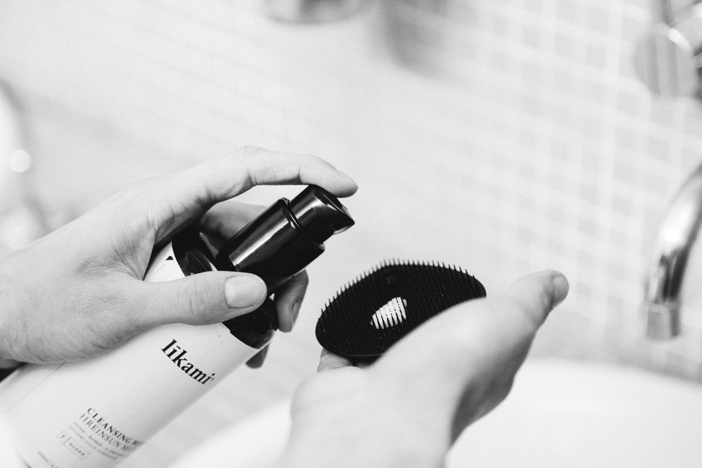 Een vrouw gebruikt de reinigingsmelk en spuit product op de reinigingsborstel om het gezicht te behandelen. Ze heeft de producten in haar handen boven een lavabo in de badkamer. Dit is een zwart-wit foto.